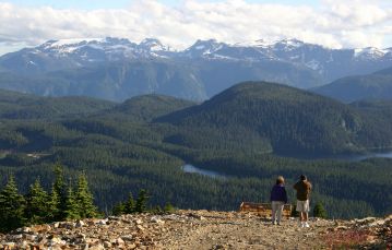 Two adults stand at view lookout point facing towards a forested valley below several lakes dot the valley there are mountains in the background and a cloudy sky Comox Vancouver Island BC Canada