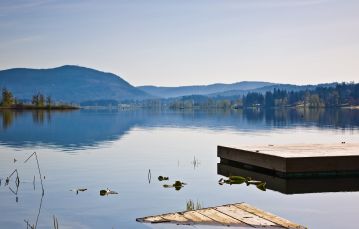 Calm blue lake waters with a wooden raft in foreground and forested mountains in background against hazy blue sky Quamichan Lake Vancouver Island BC Canada