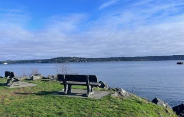 A concrete bench sits in the middle of a grassy outcropping facing towards to the ocean shoreline with blue sky Campbell River Vancouver Island BC Canada