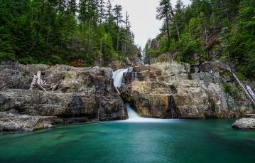 A waterfall runs down the center of two rocky shorelines and flows down into a large pool of calm blue green water pine trees are in the background against a grey sky Campbell River Vancouver Island BC Canada