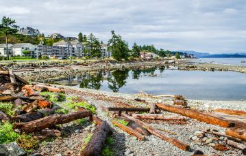 Pool of calm blue ocean water in center surrounded by shoreline dotted with driftwood residential properties re in the background set against a cloudy sky Campbell River Vancouver Island BC Canada.