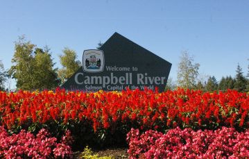Red and pink flowers line the bottom in the center a dark brown sign says welcome to Campbell River in white letters green trees behind and blue sky in back Vancouver Island BC Canada.