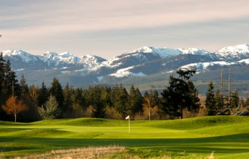 Golf course with bright green grass and one white flag in front in the center of the image with a line of green pine trees behind and snow capped mountains in the background Comox Vancouver Island BC Canada