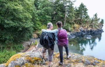 Back of male and female adults one with black backpack one with purple backpack standing atop rocky outcropping looking towards lagoon waters with pine trees along shoreline Vancouver Island Canada