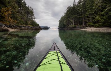 Green kayak tip pointing towards inlet with forested shoreline on either side against grey cloudy sky Vancouver Island Canada. 