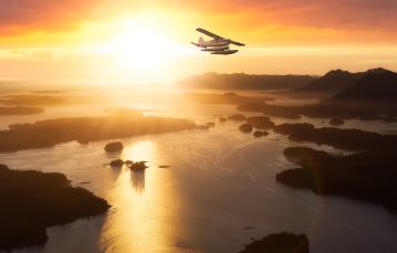 Small white Seaplane flying against bright yellow sky with gulf island below dotting ocean Vancouver Island BC Canada.