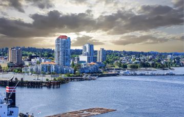 Log boom floating though blue ocean water against urban shoreline with apartment buildings and treed foothills in backdrop cloudy sky in Nanaimo Vancouver Island BC Canada.