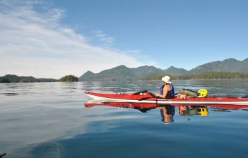 Solo kayaker cruising calm waters looking towards mountain range and blue skies of central Vancouver Island, British Columbia.
