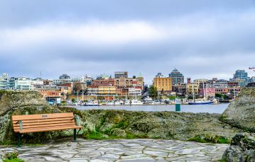 Park bench in foreground near ocean inlet with downtown Victoria buildings in background with blue sky