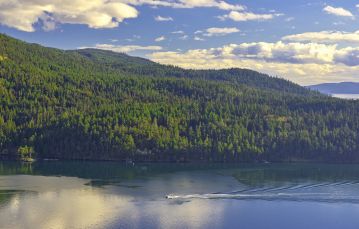Small white boat travels through calm blue waters with forested hillside in background against blue sky dotted with white clouds Salt Spring Vancouver Island BC Canada.