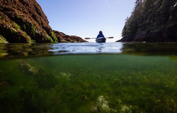 Back of kayaker floating next to rocky shoreline image split between above and below water Vancouver Island BC Canada