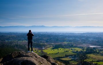Back of man in foreground standing on mount Doug in Saanich while overlooking green valley with mountain range and blue sky in background